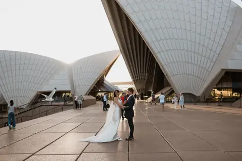 A newly married couple standing beneath the sails of the Sydney Opera House.